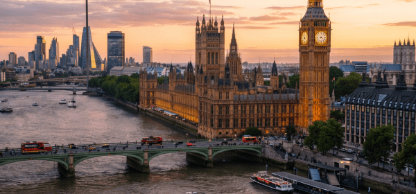 Palace of Westminster and Big Ben by the River Thames at sunset with boats and red double-decker buses on a bridge